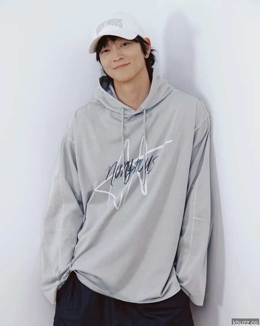 Kang Dong-won in a photoshoot still — gray hoodie, white cap, standing against a white wall